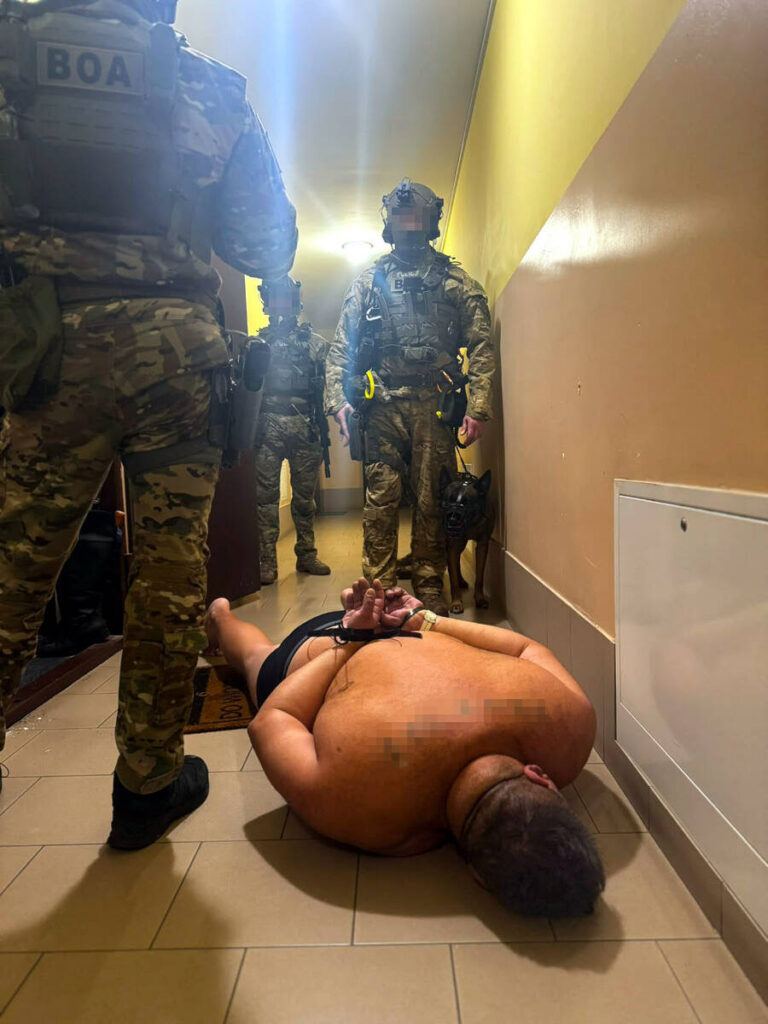 Grupa uzbrojonych funkcjonariuszy w kamizelkach taktycznych przeszukuje korytarz, a na podłodze leży mężczyzna z rękami związanymi z tyłu.