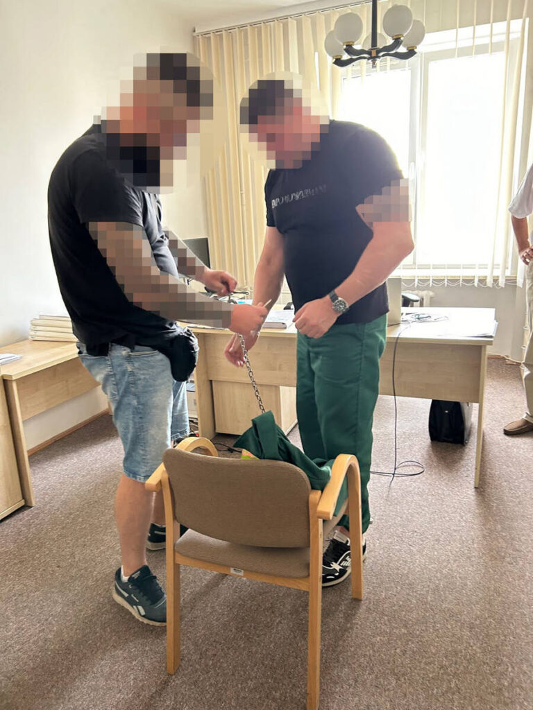 Dwaj mężczyźni w biurze zakuwają ręce jednego z nich w kajdanki, obok stoi pusta krzesło.