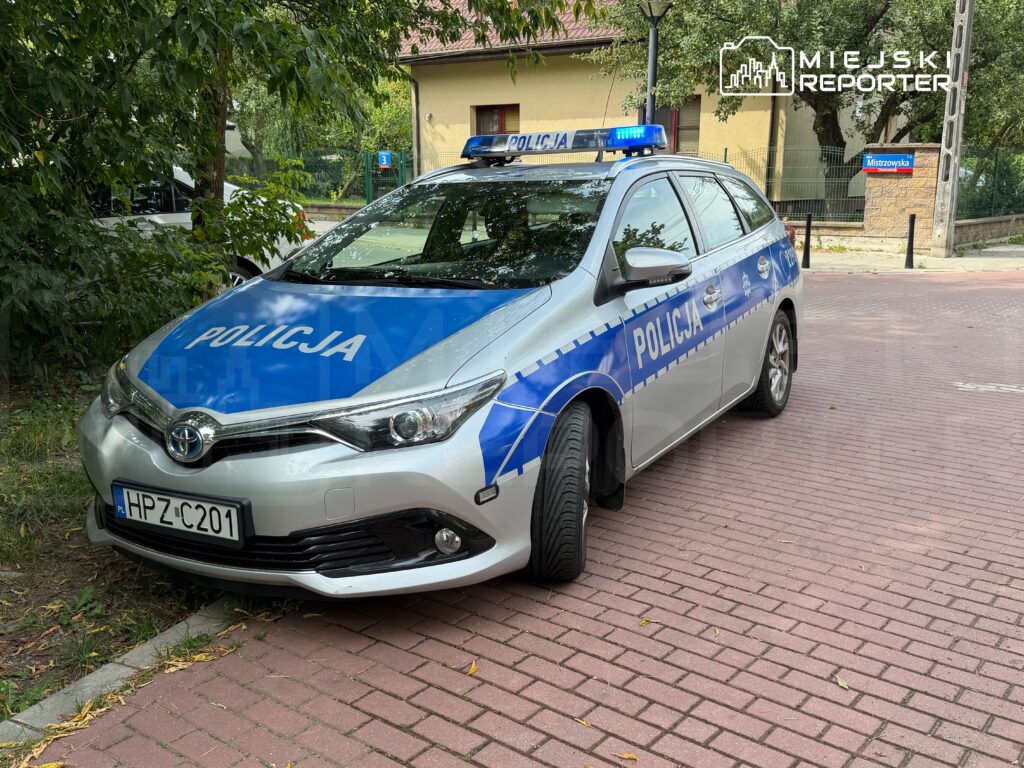 Radiowóz policyjny zaparkowany na chodniku w otoczeniu drzew i budynków mieszkalnych. Na dachu pojazdu włączone sygnały świetlne.