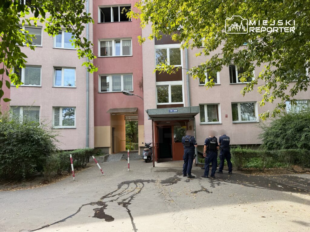 Trzech policjantów w mundurach przechodzi obok wejścia do bloku mieszkalnego, w otoczeniu drzew i krzewów.