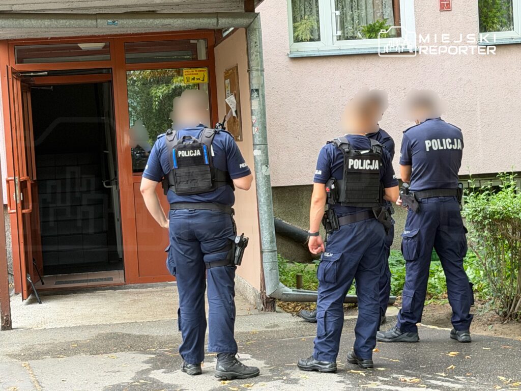 Trzech policjantów w mundurach rozmawia przed wejściem do budynku mieszkalnego.