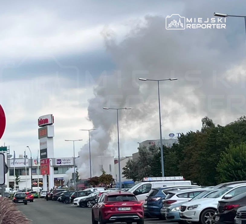 Gęsty, biały dym unoszący się z budynku w tle, otoczony przez parking z zaparkowanymi samochodami.