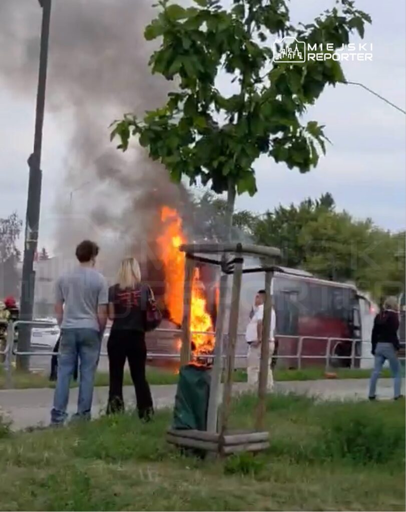 Grupa osób obserwuje płonący autobus, z którego wydobywa się dym, w miejskim otoczeniu.