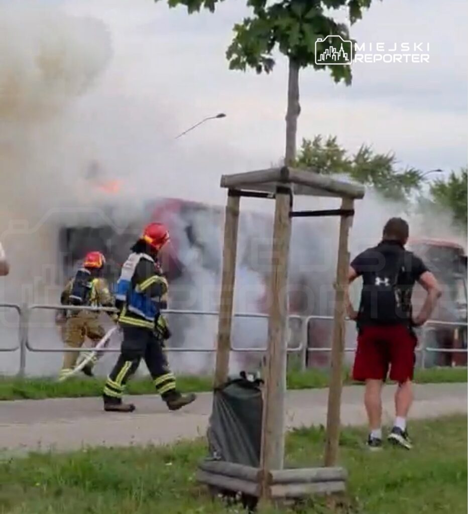 Strażacy w mundurach gaszą płonący pojazd, otoczeni przez gęsty dym, w miejskim otoczeniu.