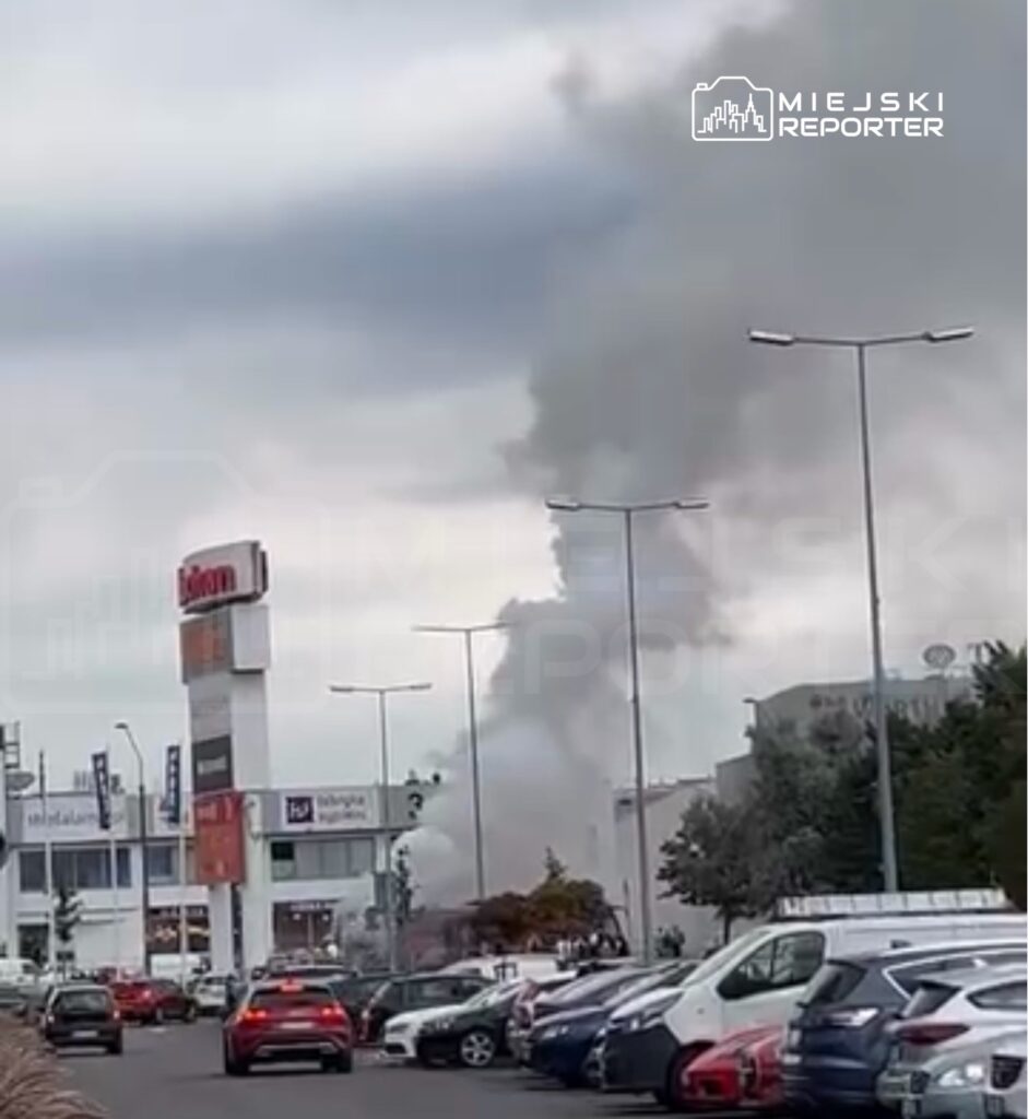Gęsty dym unoszący się nad nowoczesnym kompleksem handlowym, w tle widoczne samochody na parkingu.