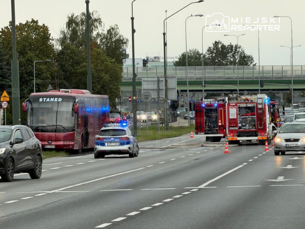 Czerwony autobus z napisem "STANED" zatrzymany na poboczu, obok radiowóz policyjny i wozy straży pożarnej z włączonymi sygnałami świetlnymi.