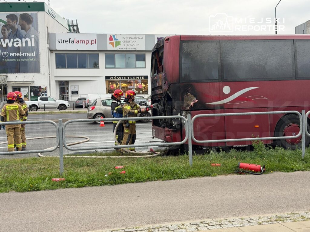 Strażacy gaszą pożar autobusu na drodze, obok stoją uszkodzone pojazdy i gaśnice. W tle widoczne budynki.