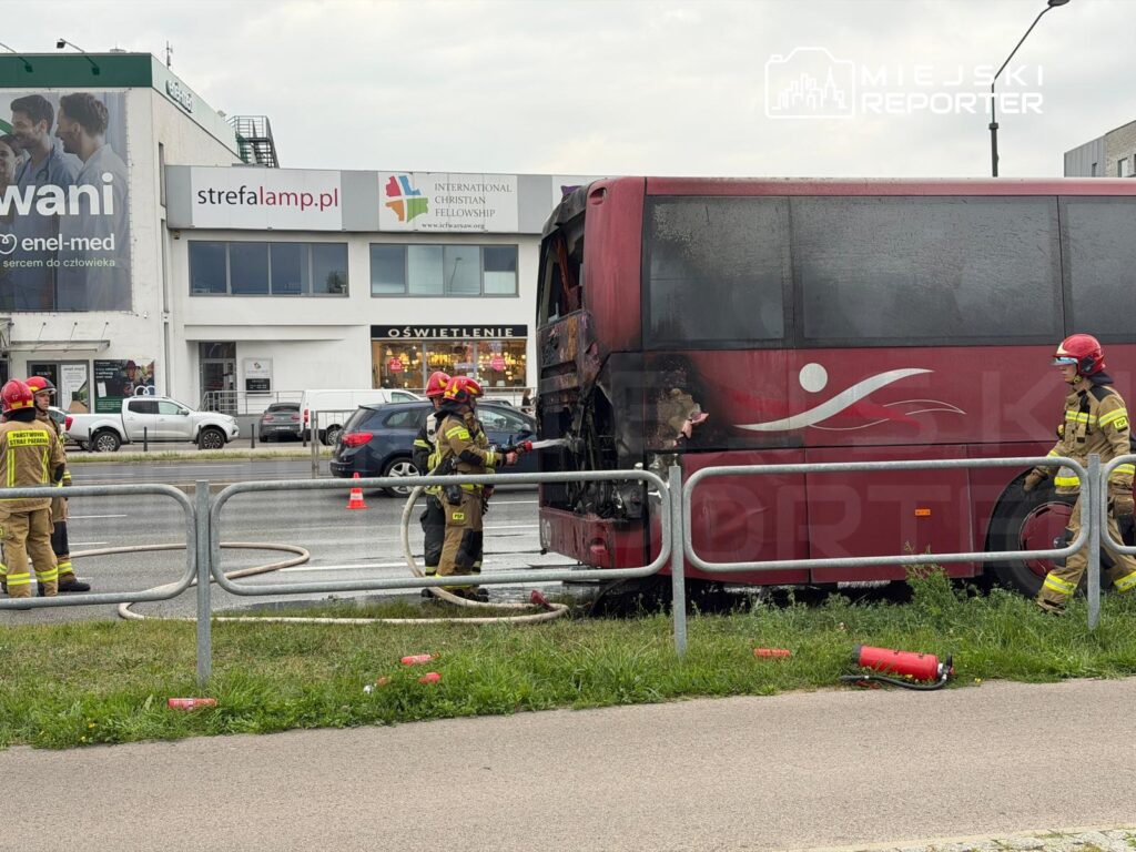 Grupa strażaków w uniformach gasi pożar tyłu autobusu na ruchliwej ulicy, obok widoczne budynki i pojazdy.