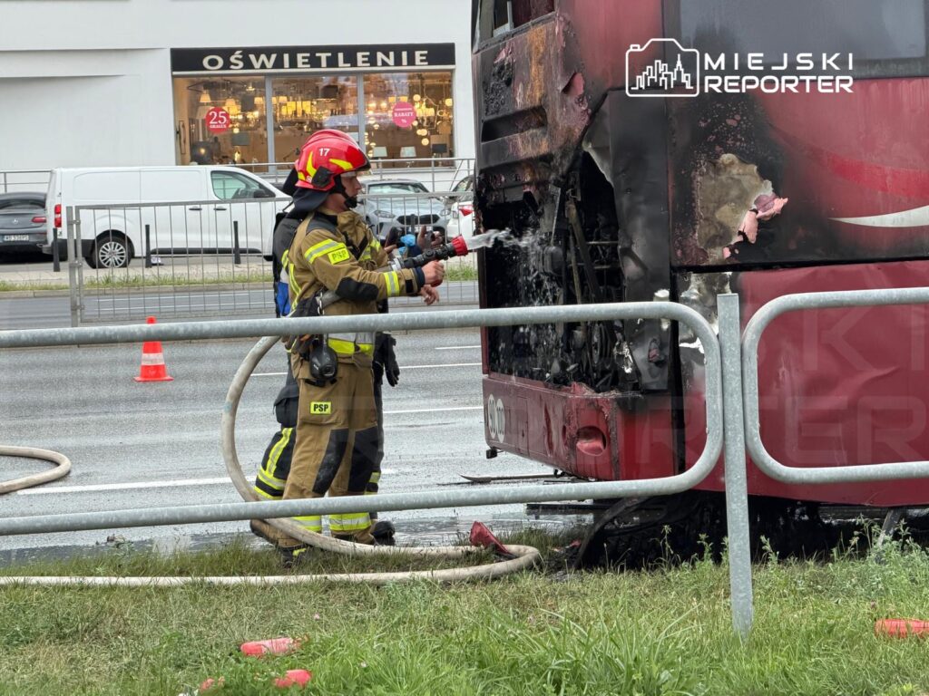 Strażak w mundurze gaśniczym używa węża strażackiego do gaszenia ognia w spalonym autobusie na ulicy.