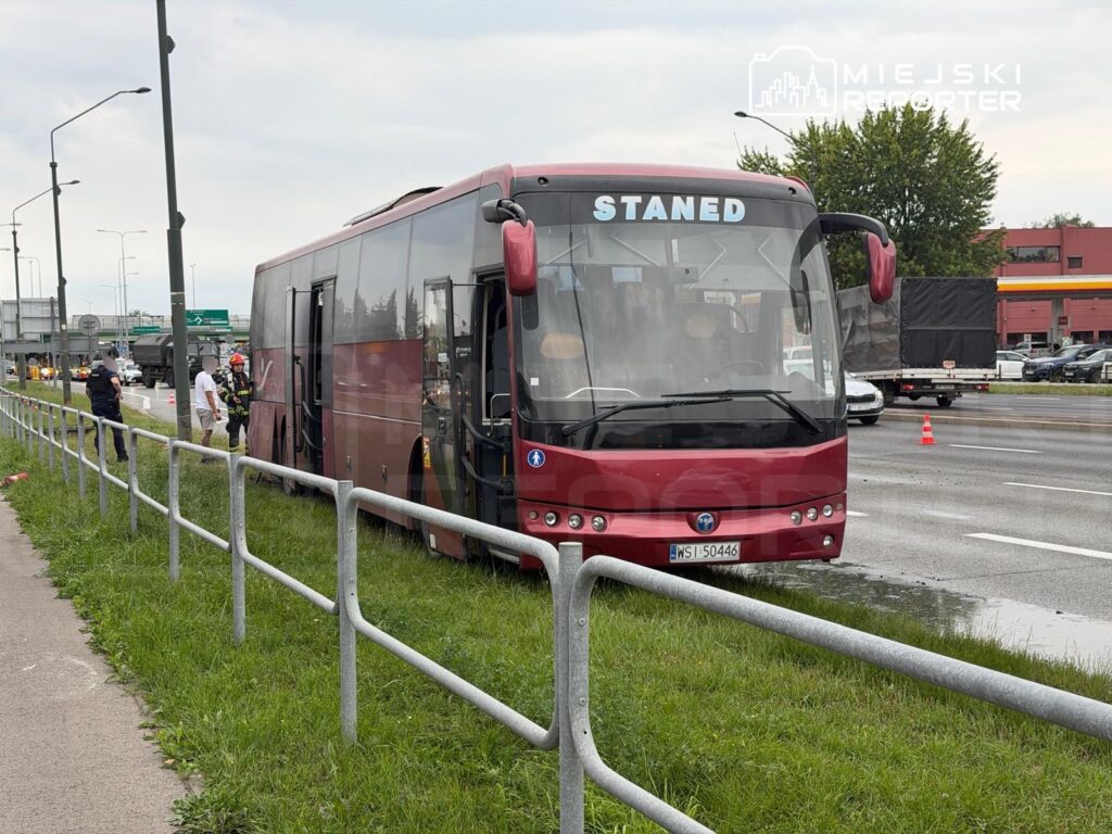 Czerwony autobus stoi na poboczu drogi, obok grupy strażaków i policjantów, w tle ruch drogowy.