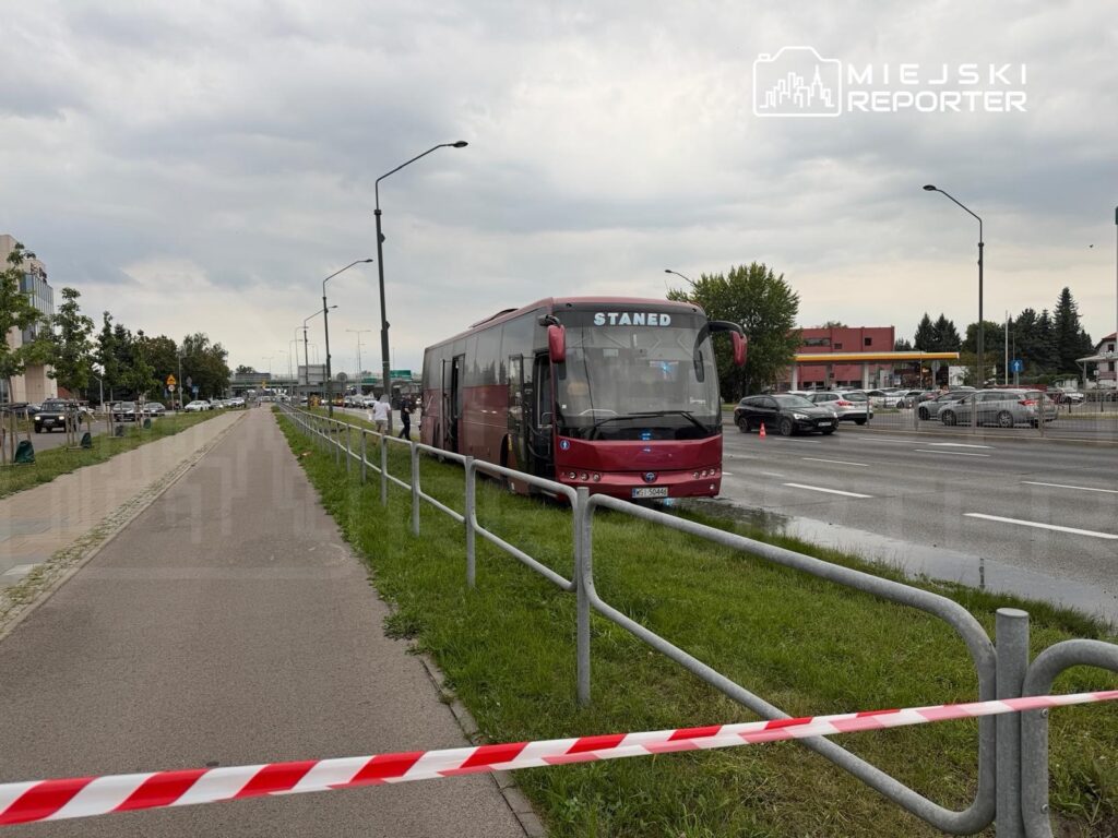 Czerwony autobus stoi przy drodze, a obok niego zabezpieczenie w postaci taśmy ostrzegawczej. W tle widoczne są samochody i budynki.