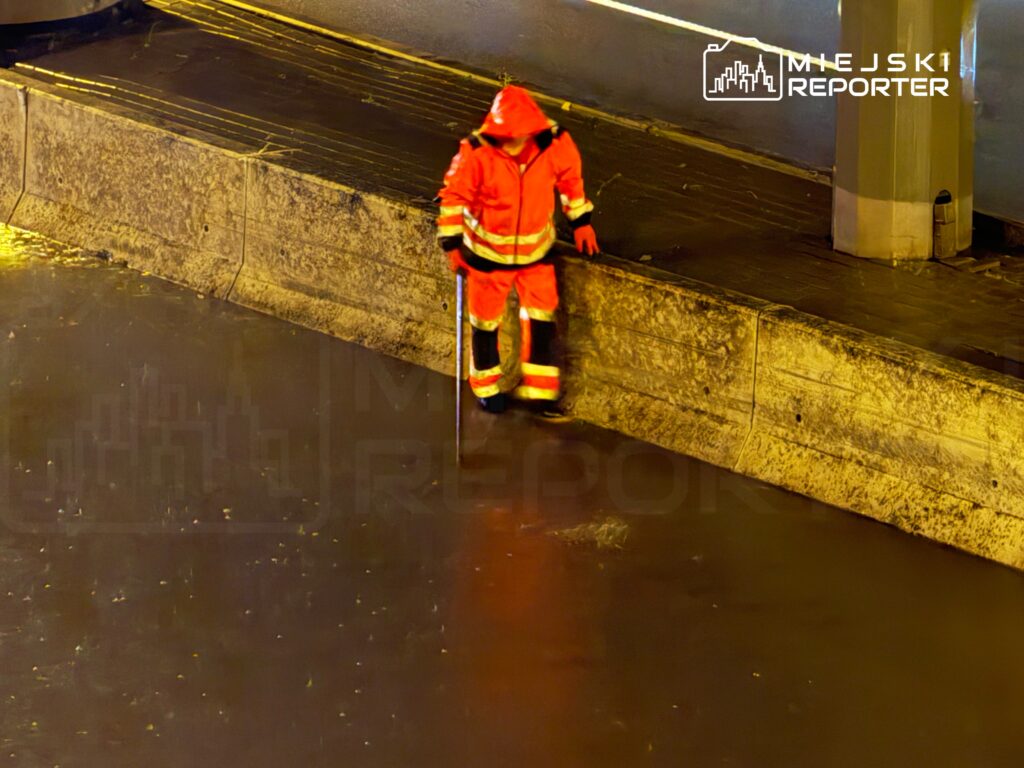 Ratownik w pomarańczowym uniformie sprawdza poziom wody na zalanej ulicy nocą.