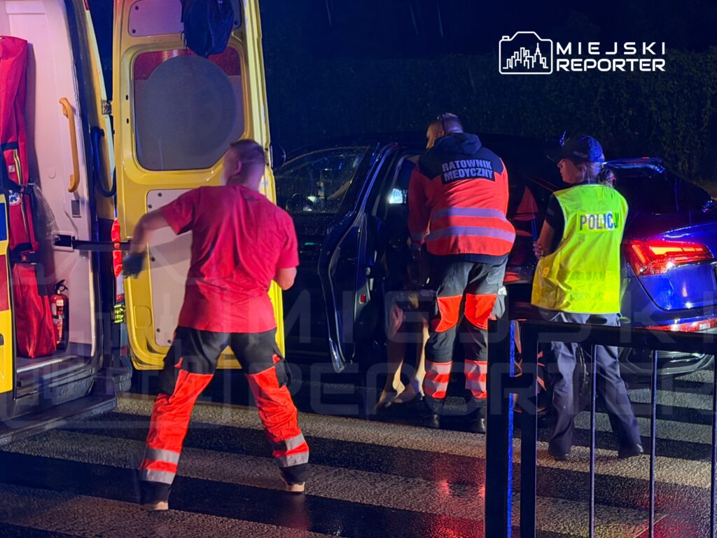 Ratownicy medyczni udzielają pomocy osobie siedzącej w samochodzie, obok stoi policjant. W tle widać ambulans.