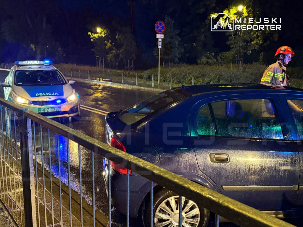 Policjant w hełmie rozmawia z kierowcą na mokrej ulicy, obok zaparkowany radiowóz z włączonymi sygnałami świetlnymi.