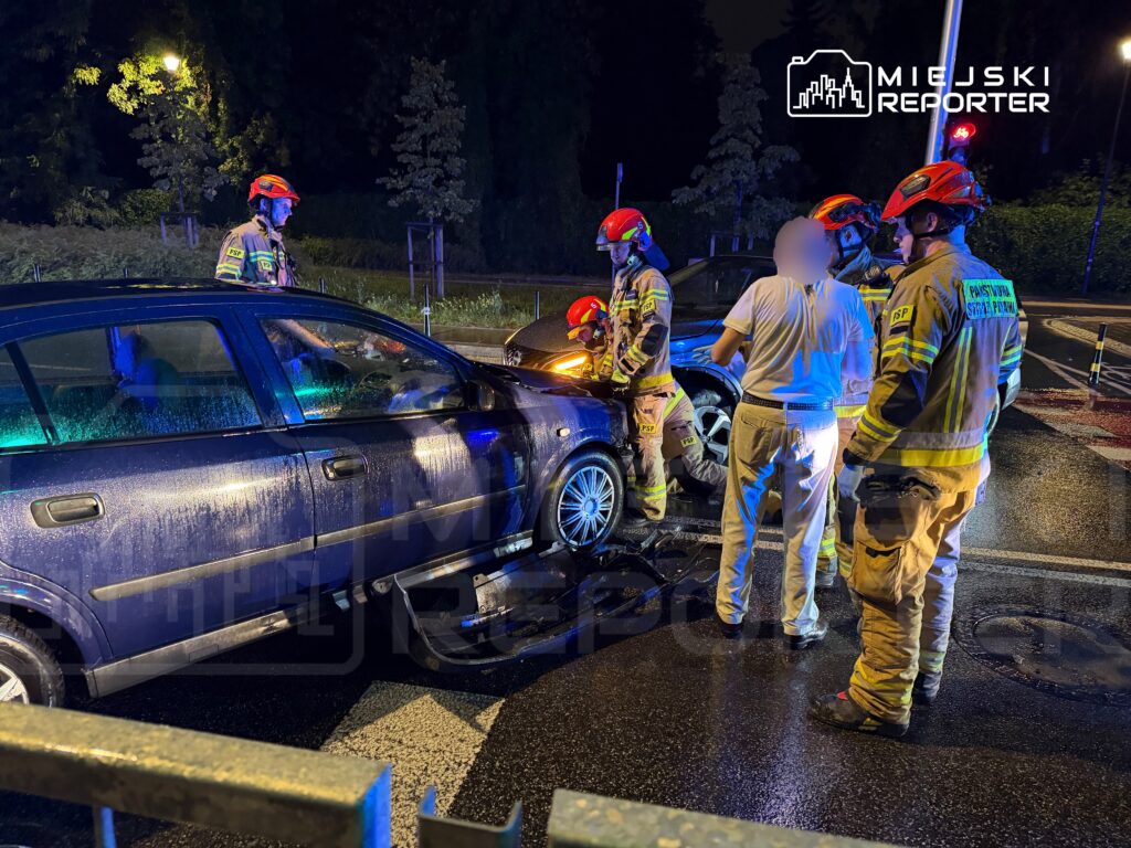 Czterech ratowników w mundurach rozmawia z kierowcą przy uszkodzonym samochodzie na nocnej ulicy.