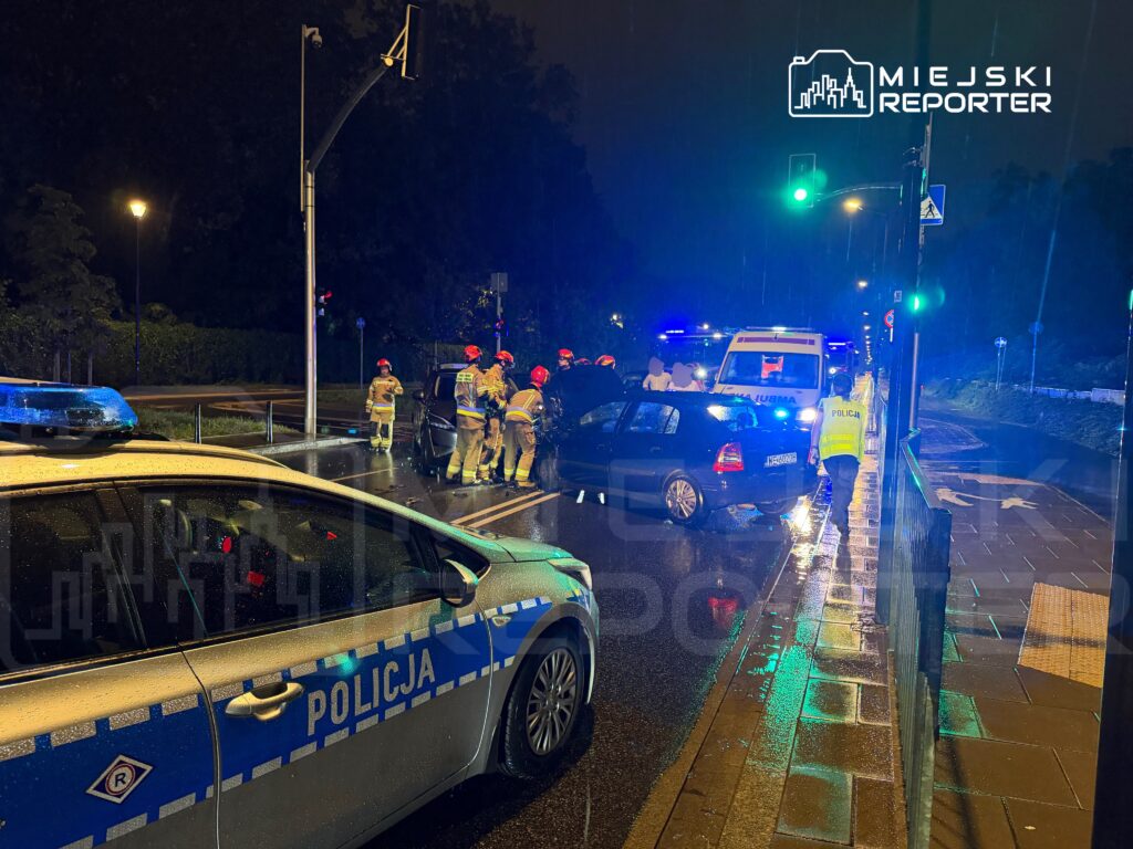 Grupa strażaków i policjantów udziela pomocy po wypadku drogowym na deszczowej ulicy, obok pojazdów służb.