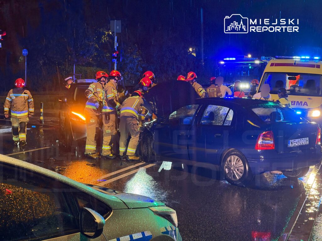 Grupa strażaków pracuje nad usunięciem uszkodzonego samochodu po wypadku drogowym w deszczowej nocy.