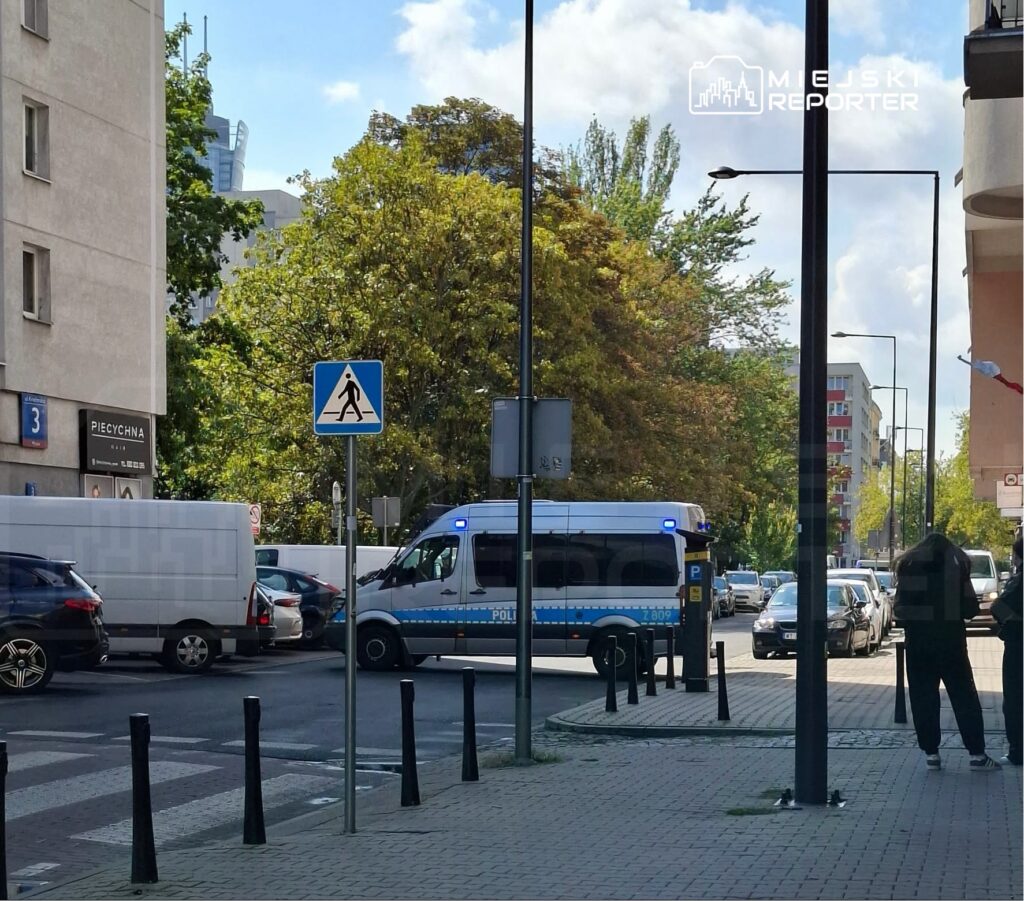 Policyjny van z włączonymi sygnalizatorami świetlnymi zatrzymany na skrzyżowaniu, obok przejścia dla pieszych.