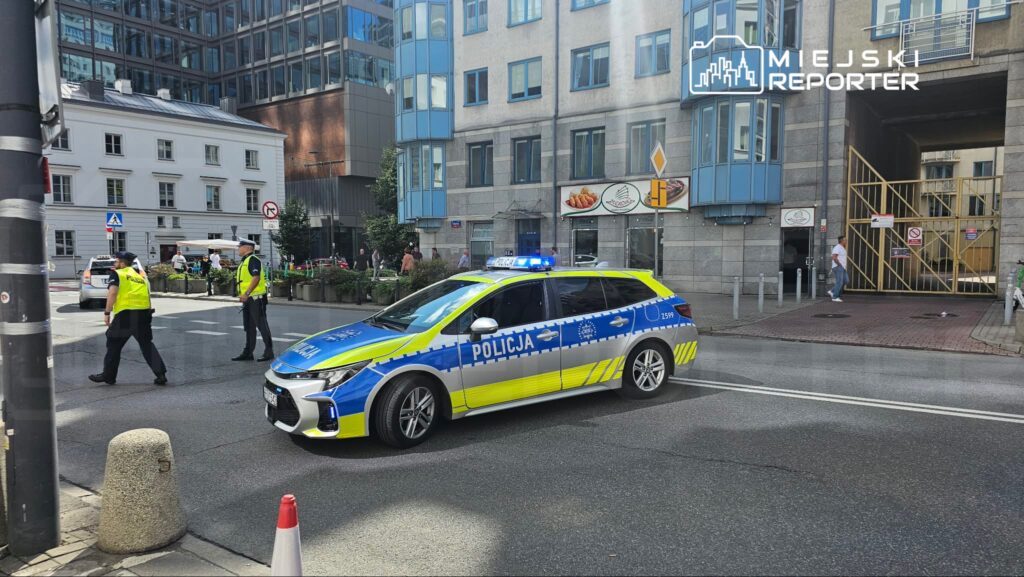 Radiowóz policyjny z niebieskimi światłami blokuje ulicę, a policjanci w kamizelkach kierują ruchem w miejskim otoczeniu.