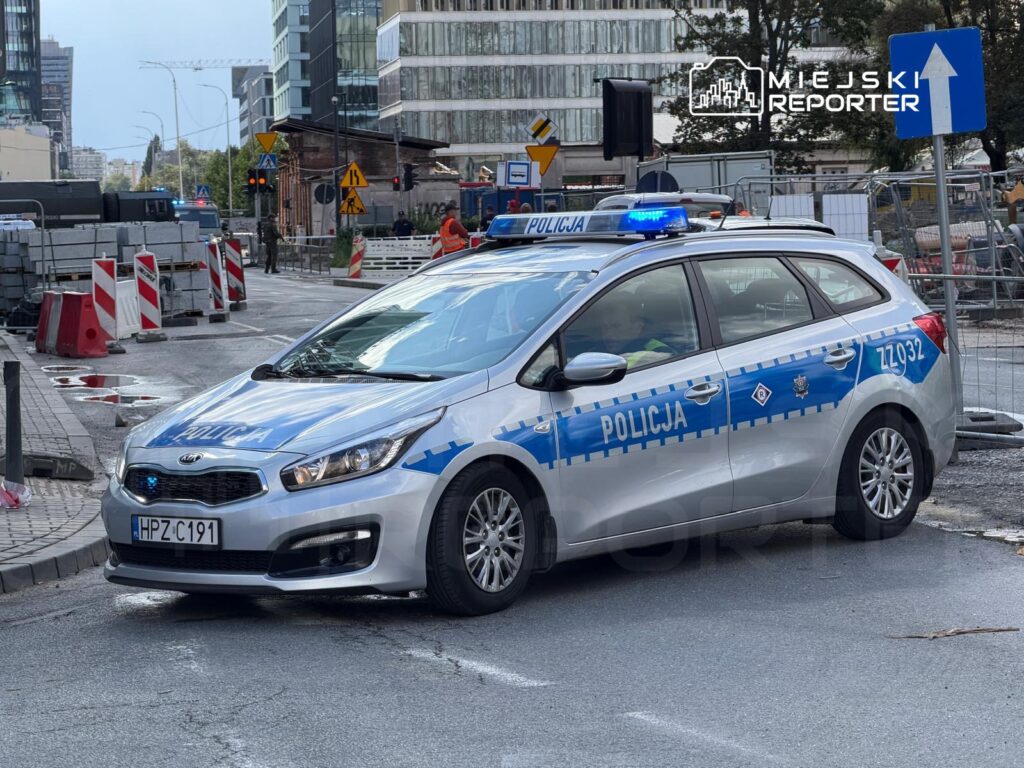 Policyjny radiowóz z włączonymi sygnałami świetlnymi stoi na zablokowanej ulicy, otoczony przez sprzęt budowlany.