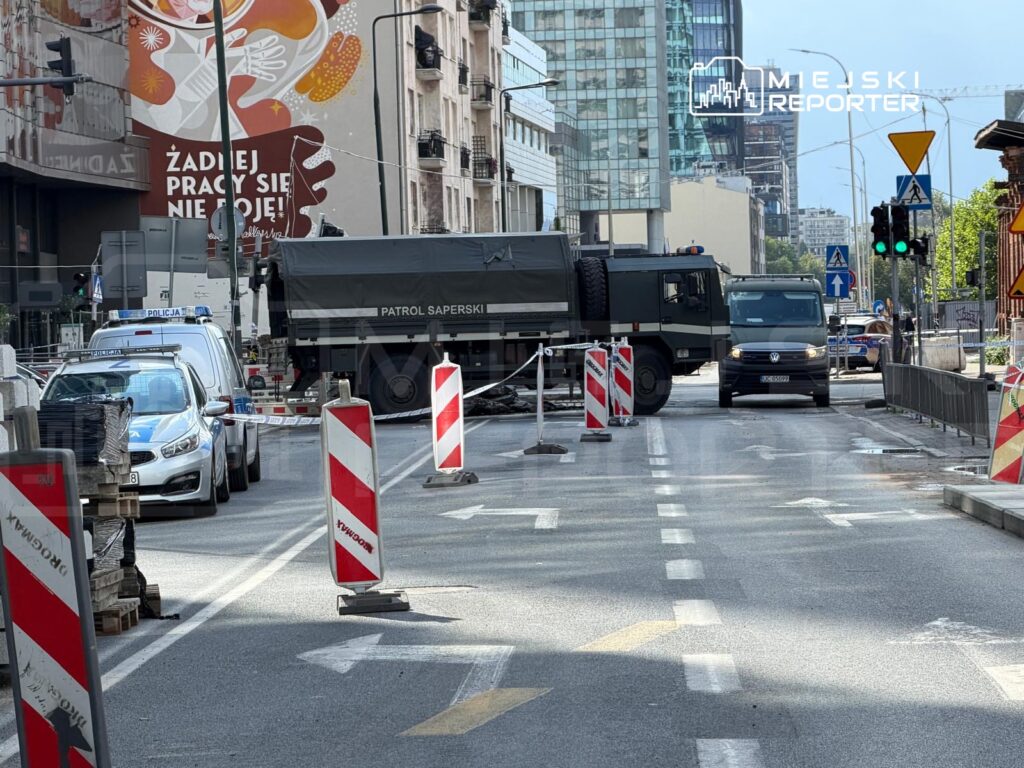 Na ulicy widać policyjny samochód oraz pojazd wojskowy, a droga jest zablokowana przez bariery budowlane.