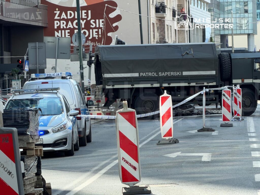 Radiowóz policyjny oraz pojazd z napisem "PATROL SAPERSKI" blokują ulicę, otoczony taśmą zabezpieczającą i słupkami. W tle widoczny mural z hasłem "ŻADNEJ PRACY SIĘ NIE BOJĘ!".