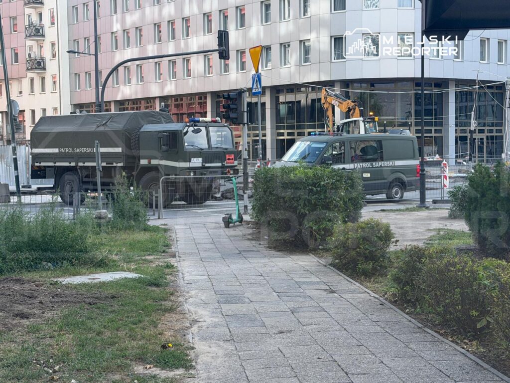 Na skrzyżowaniu stoją pojazdy z napisem "PATROL SAPERSKI", obok widać maszynę budowlaną oraz zieleń.