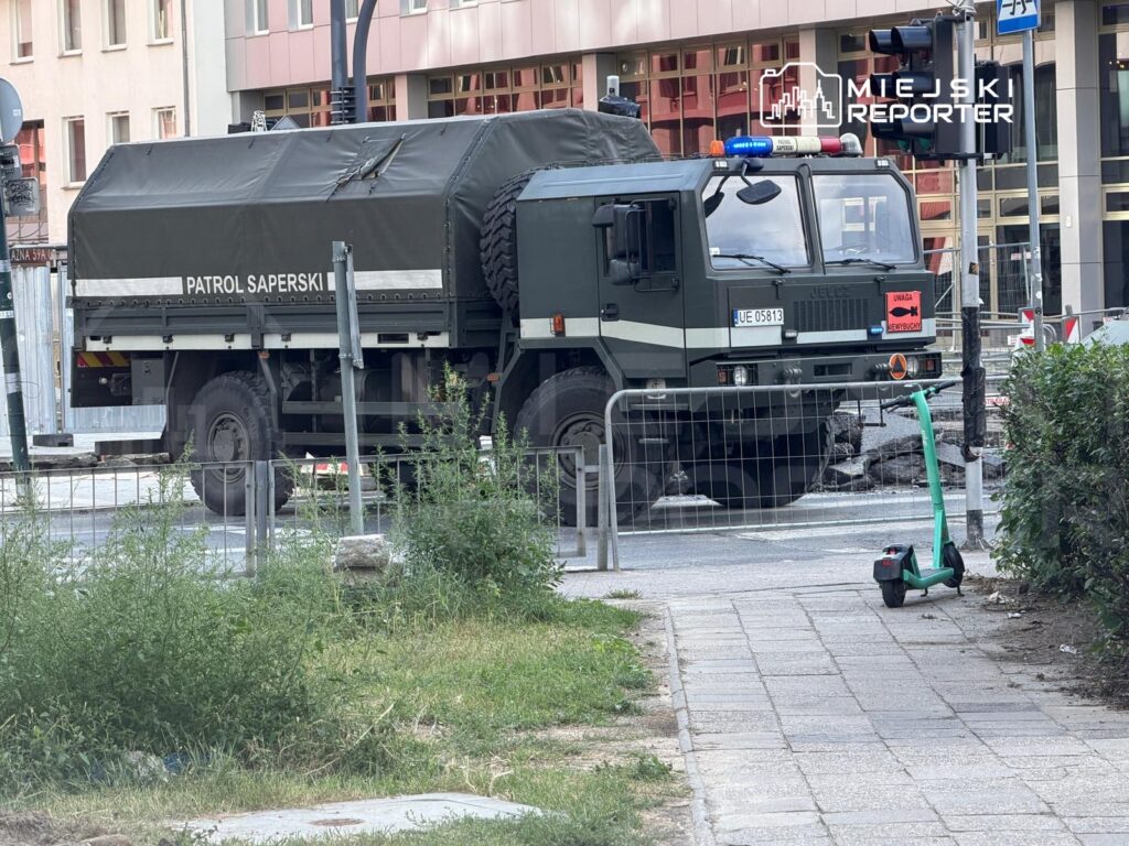 Militarny pojazd z napisem "PATROL SAPERSKI" zatrzymany na ulicy obok zielonej hulajnogi, w miejskim otoczeniu.