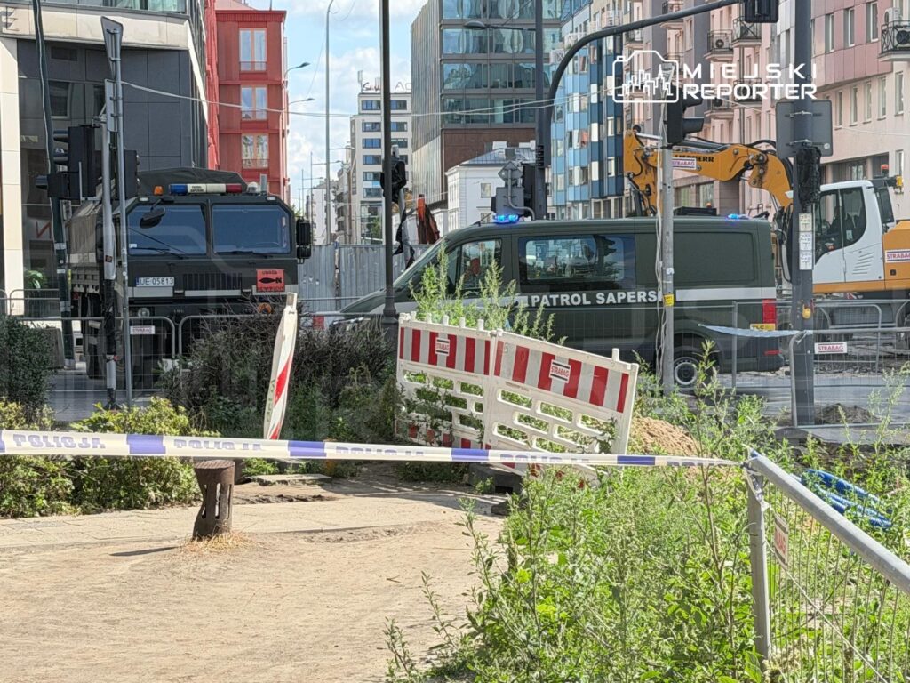 Na ulicy stoją dwa pojazdy służbowe: ciężarówka z sygnalizacją świetlną i furgon z napisem „PATROL SAPERSKI”, otoczone barierkami.