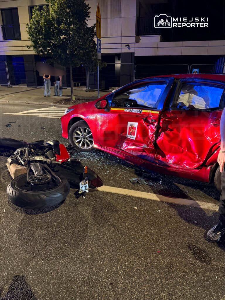 Czerwony samochód osobowy z wgniecioną stroną stoi na ulicy obok przewróconego motocykla, w tle widać ludzi.