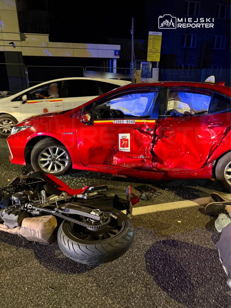 Czerwony samochód osobowy z uszkodzoną boczną częścią stoi na drodze, obok leży przewrócony motocykl.