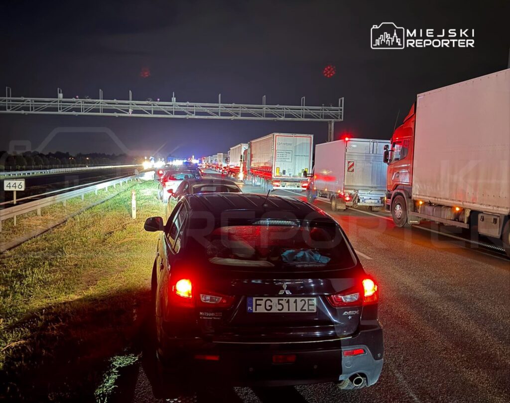Samochody stoją w długim korku na autostradzie w nocy, z widocznymi światłami i ciężarówkami w tle.