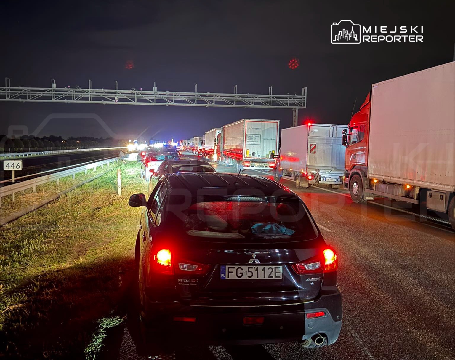Samochody stoją w długim korku na autostradzie w nocy, z widocznymi światłami i ciężarówkami w tle.