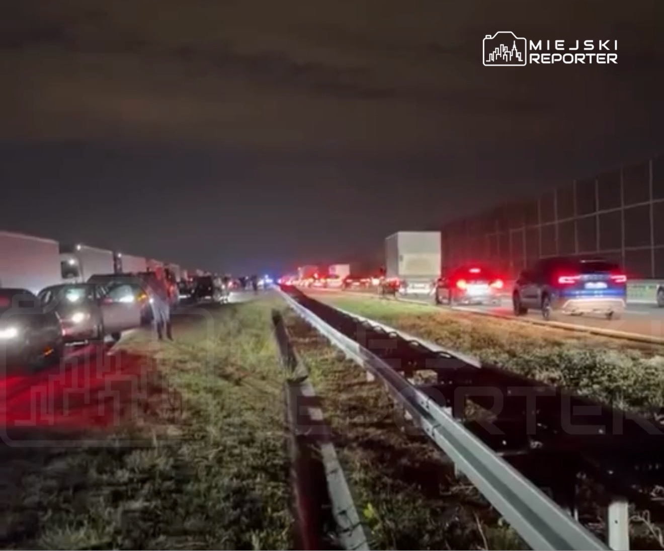 Ruch samochodowy utknął w korku na drodze, w nocy, z widocznymi światłami pojazdów i ludzi stojących obok.