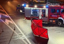 Na drodze widoczny jest wóz strażacki z otwartą zabudową, obok znajduje się policjant zabezpieczający miejsce zdarzenia.