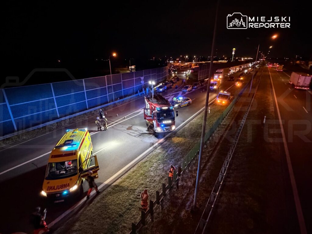 Na drodze w nocy interweniują służby ratunkowe, w tym ambulans i wóz strażacki, otoczone pojazdami.