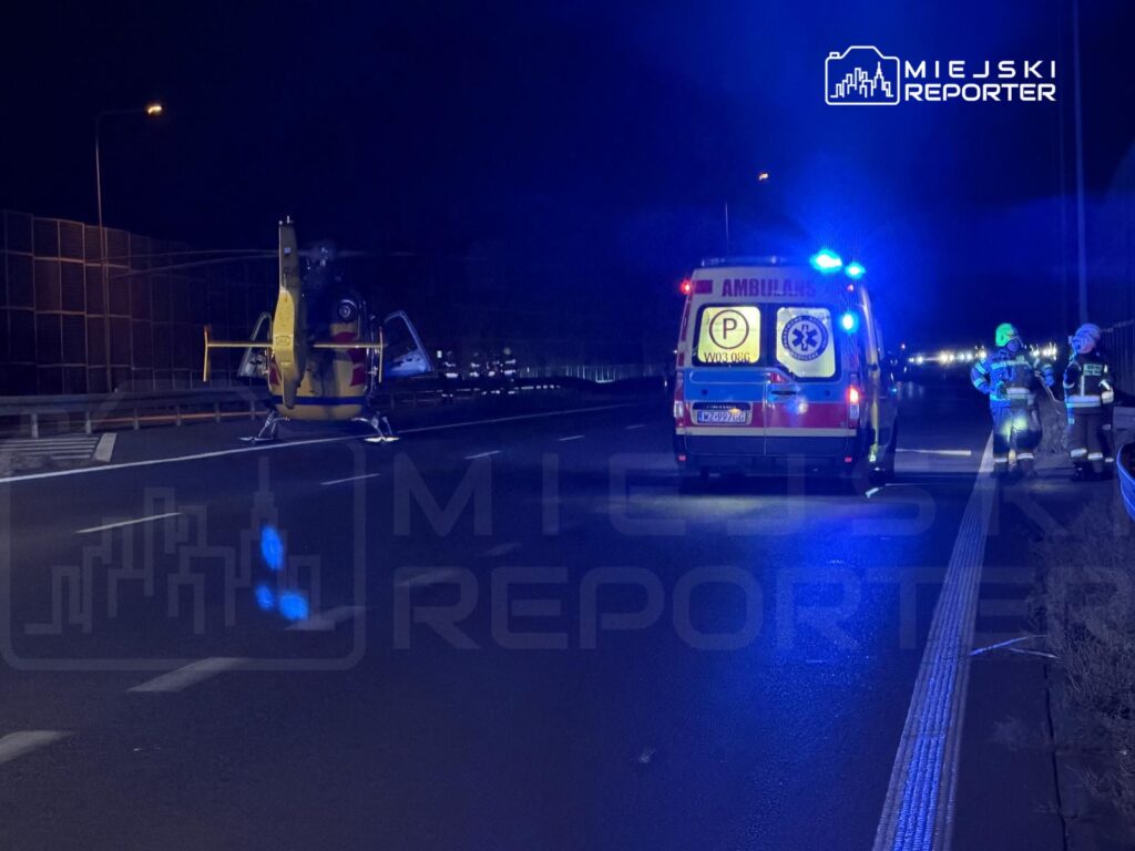 Ambulans z włączonymi sygnałami świetlnymi zatrzymany na drodze, obok lądowisko dla helikoptera ratunkowego.