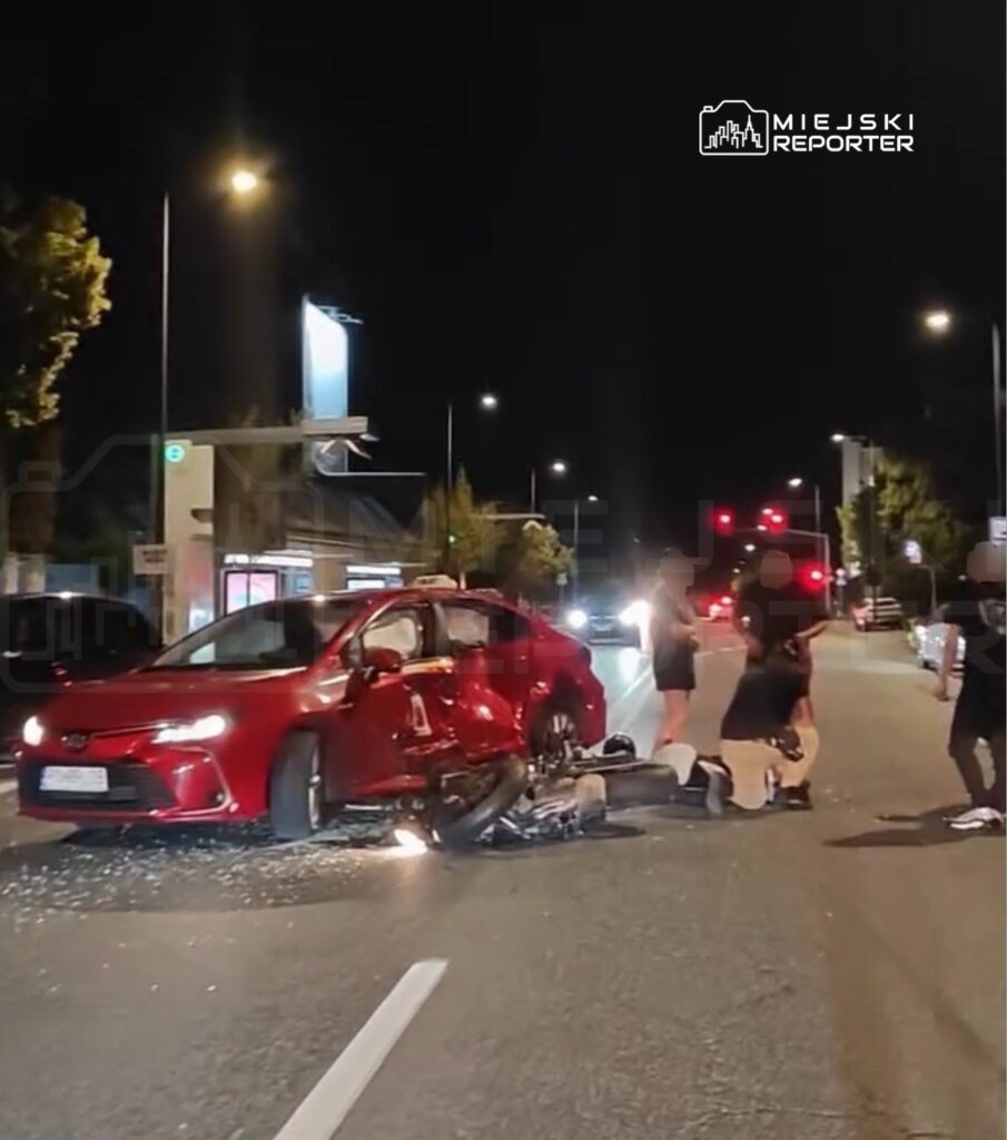 Czerwony samochód osobowy stoi na drodze, obok leży motocykl, a kilka osób udziela pomocy poszkodowanemu.