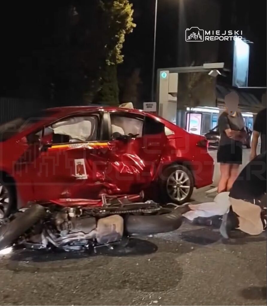 Czerwony samochód osobowy z uszkodzoną boczną częścią stoi na jezdni, obok leży przewrócony motocykl, a obok grupka ludzi.