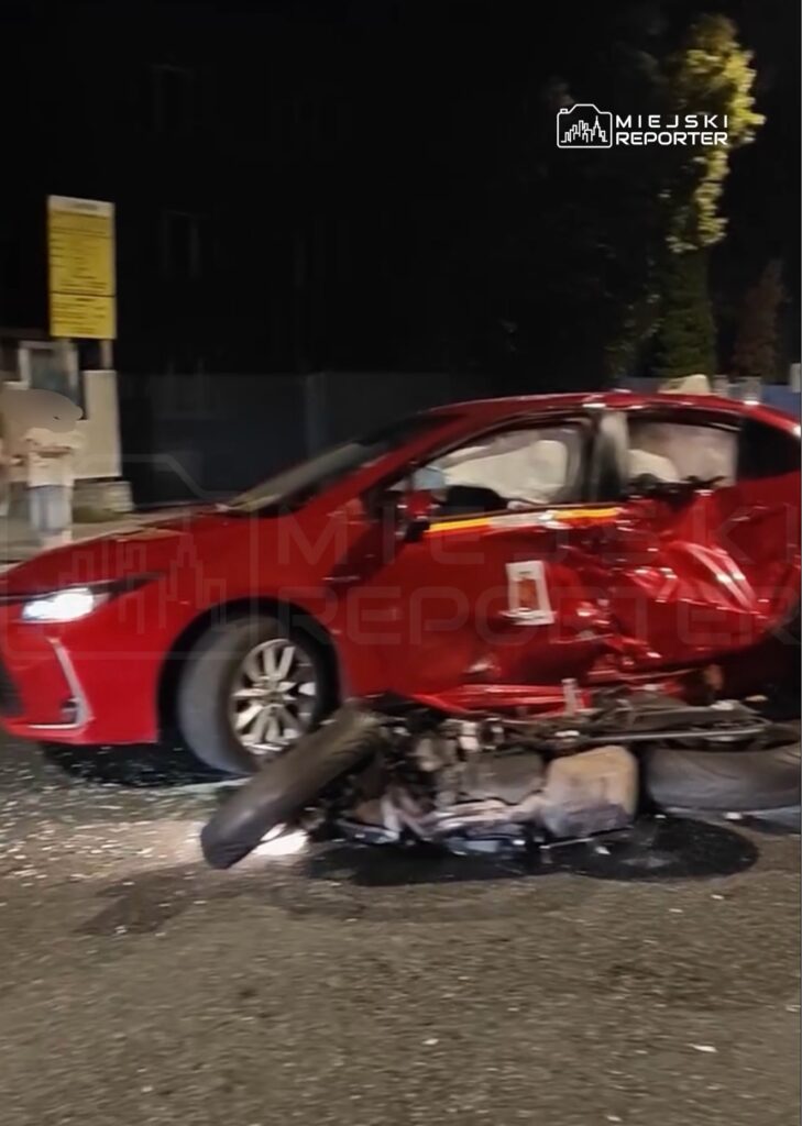 Uszkodzony czerwony samochód osobowy stoi na jezdni, obok leży oderwany silnik i koła. W tle widoczny znak drogowy.