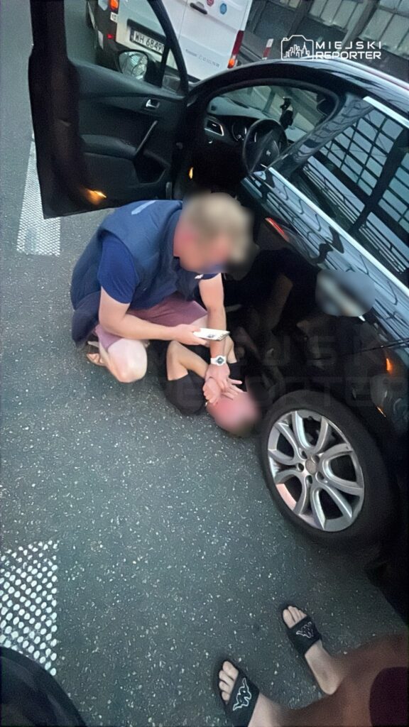 Mężczyzna w krótkich spodniach klęczy obok leżącego na jezdni mężczyzny, trzymając w ręku telefon.