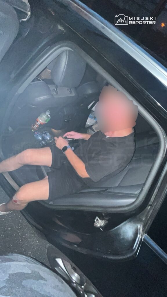 Mężczyzna w czarnym stroju siedzi na tylnej kanapie samochodu, trzymając telefon i napój.