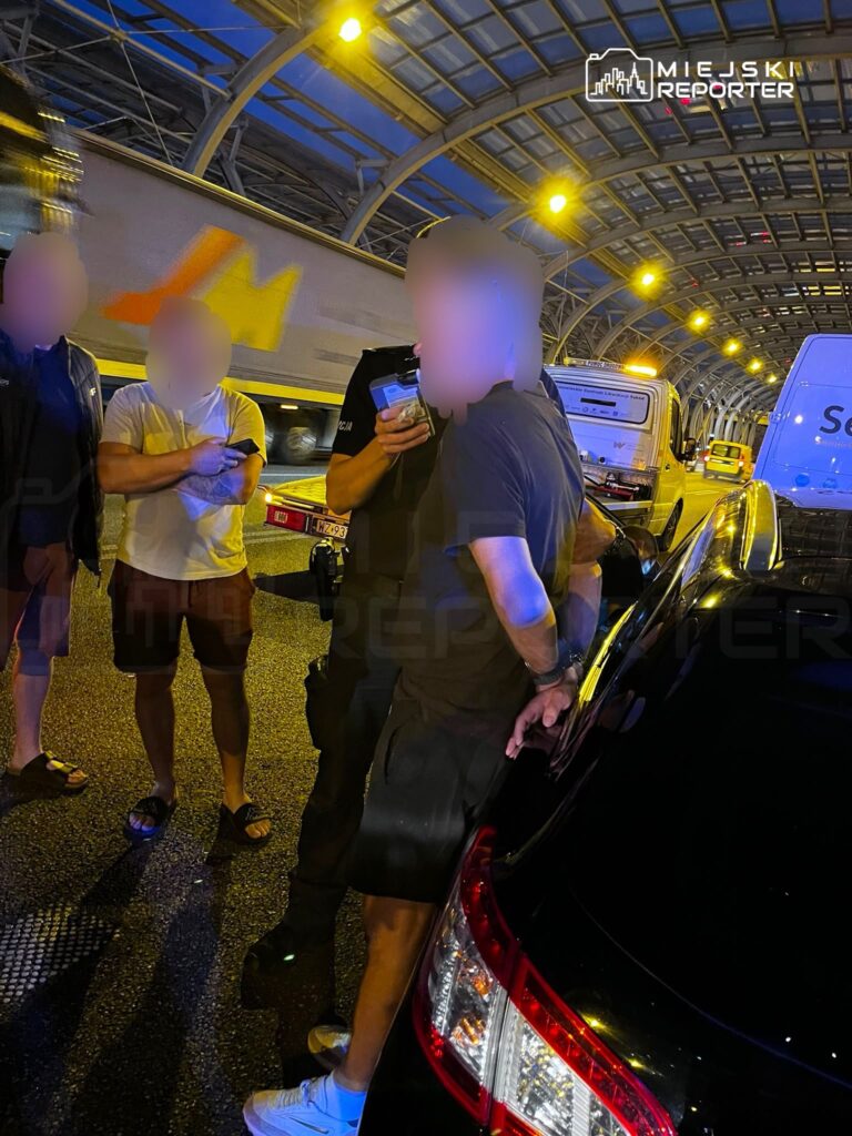 Grupa mężczyzn rozmawia z policjantem na drodze pod zadaszoną konstrukcją, w tle przejeżdżające pojazdy.