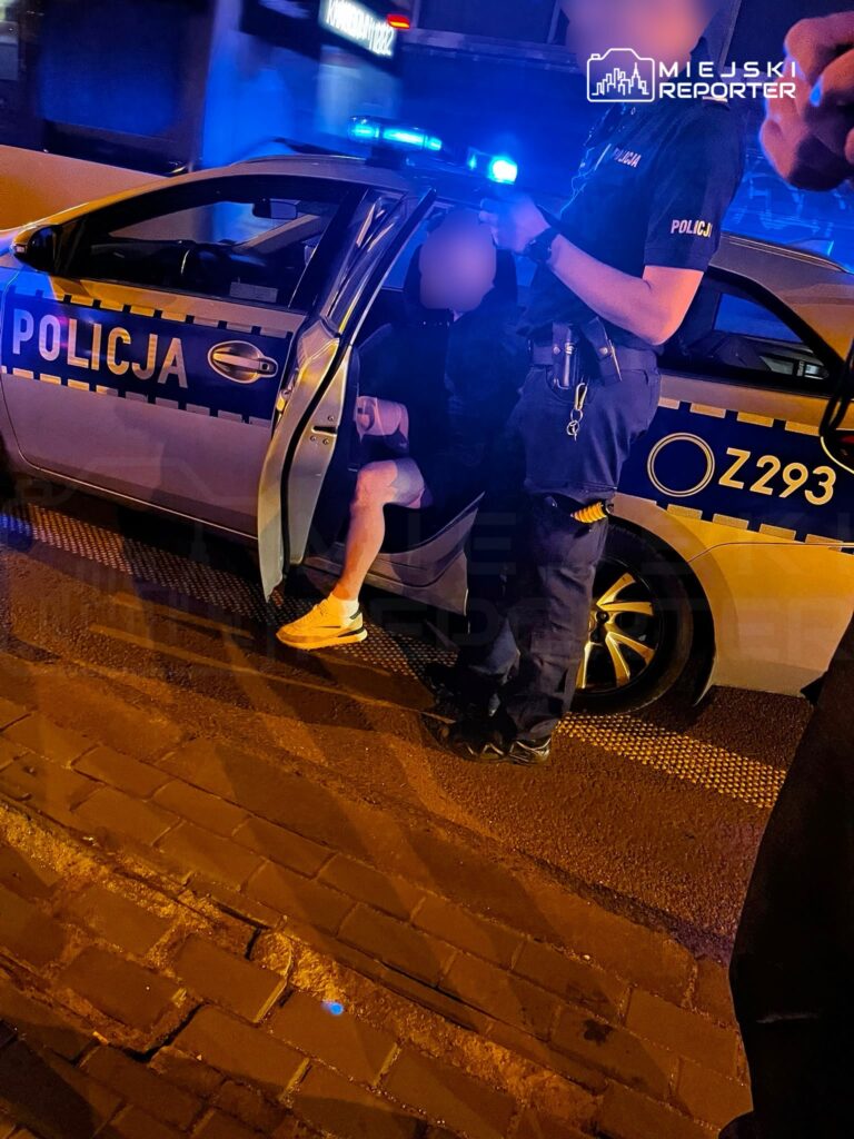 Policjant w mundurze interweniuje przy otwartych drzwiach radiowozu, zatrzymując mężczyznę w krótkich spodniach.