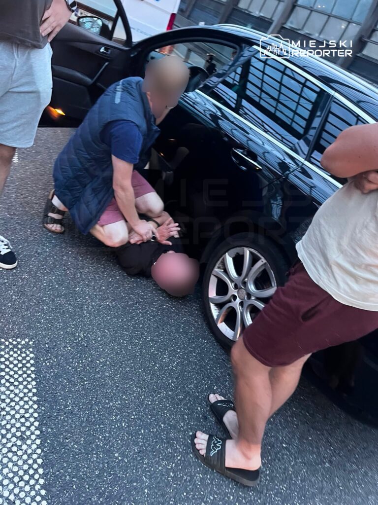 Mężczyzna w niebieskiej kamizelce przytrzymuje osobę na ziemi obok czarnego samochodu na ulicy. W tle widać innych mężczyzn.