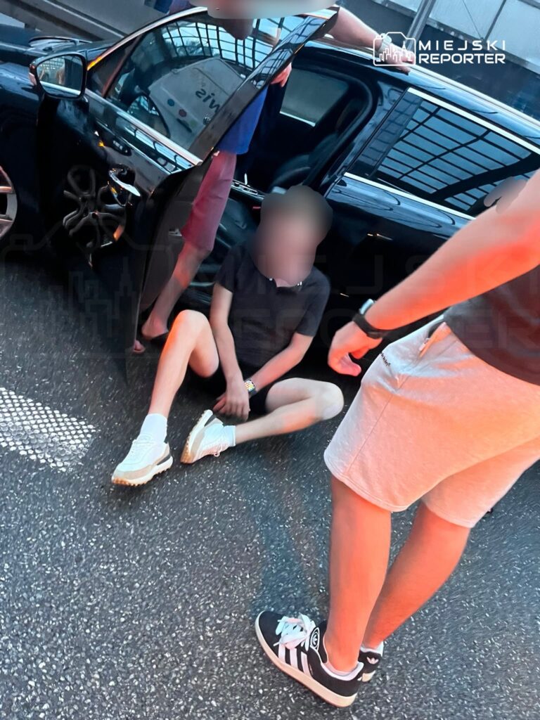 Młody mężczyzna siedzący na jezdni obok otwartego samochodu, otoczony przez dwóch innych mężczyzn.