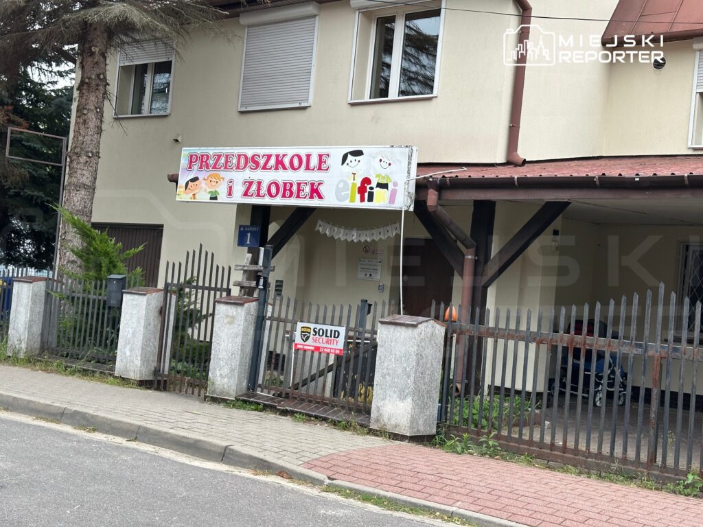 Przedszkole i żłobek z kolorowym szyldem, ogrodzone niskim płotem, w otoczeniu drzew i chodnika.