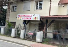 Przedszkole i żłobek z kolorowym szyldem, ogrodzone niskim płotem, w otoczeniu drzew i chodnika.
