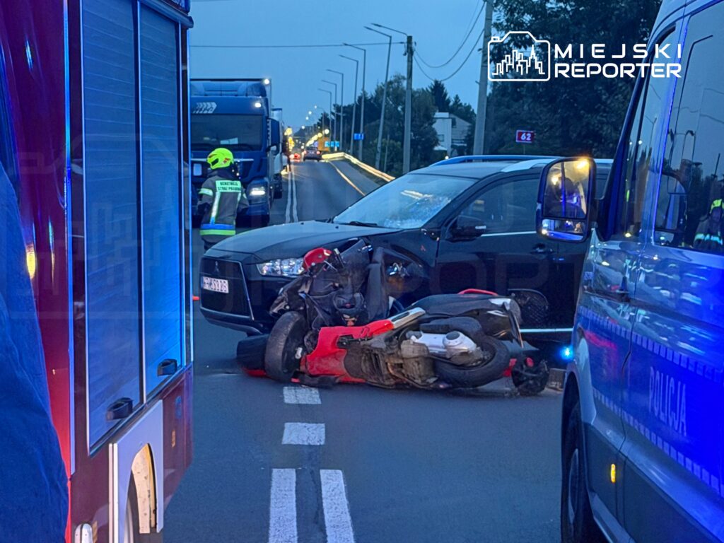 Motocykl leży na boku na drodze, obok czarnego samochodu osobowego, a policjant bada miejsce wypadku.