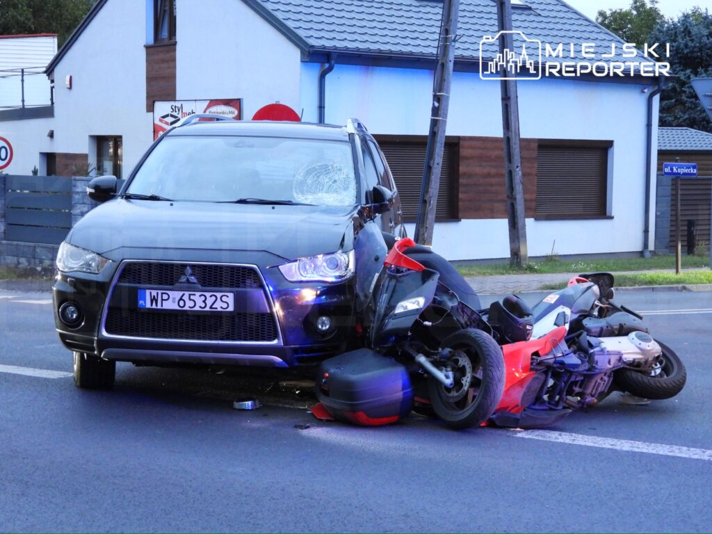 Czarny samochód osobowy z uszkodzoną szybą stoi obok przewróconego motocykla na drodze.